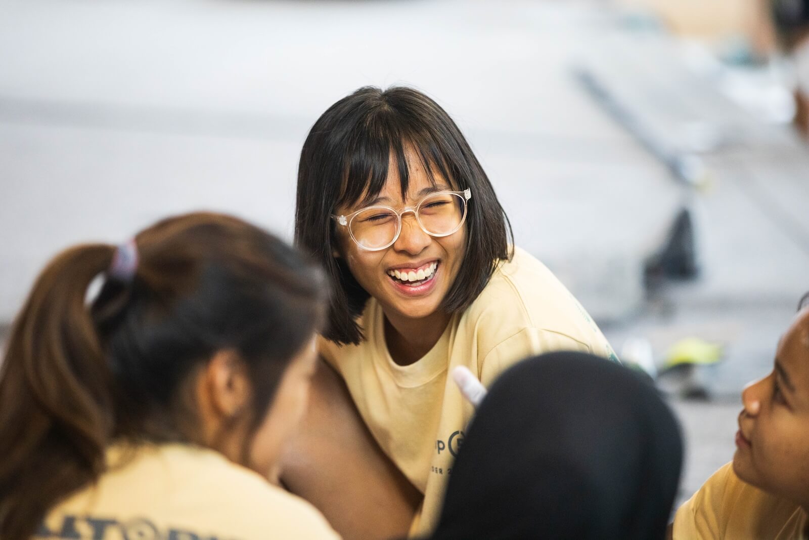 A candid medium close-up of a young woman with a short black bob and clear-framed glasses laughing heartily with her eyes squinted shut, sitting in a circle with other participants wearing matching pale yellow t-shirts at a climbing competition in Kuala Lumpur.