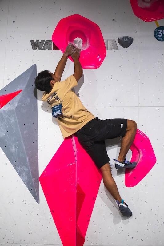 A climber in a tan t-shirt and black shorts scales an indoor bouldering wall, gripping a large, bright pink circular volume with both hands.