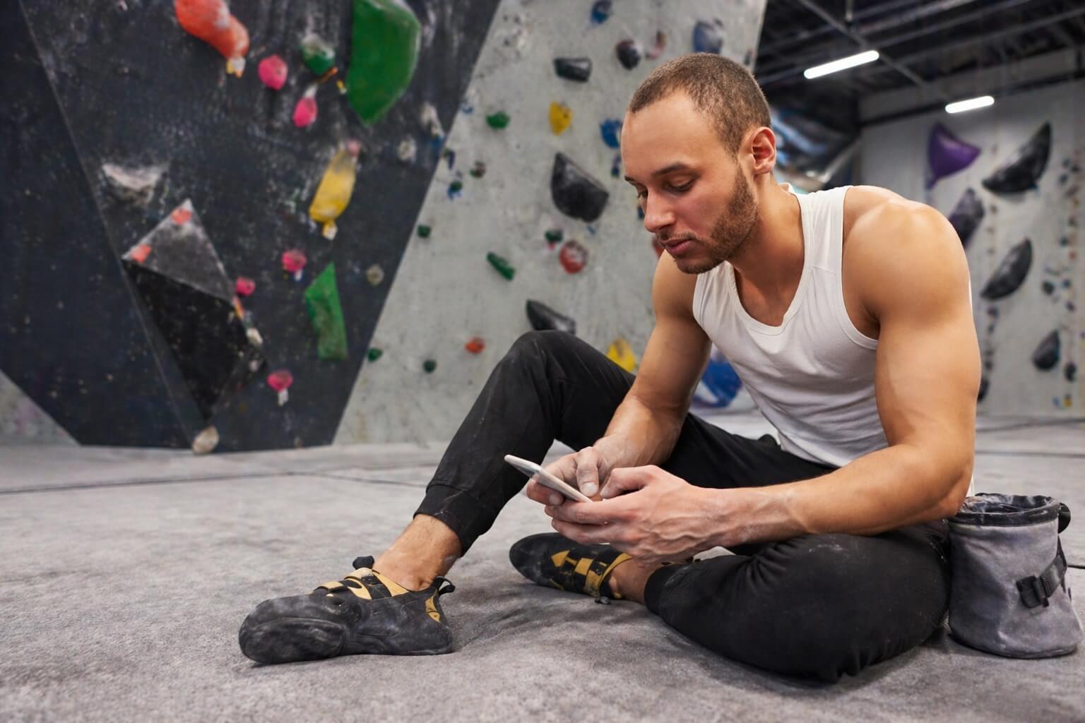 A man sits cross-legged on a climbing gym floor checking his phone next to a chalk bag.