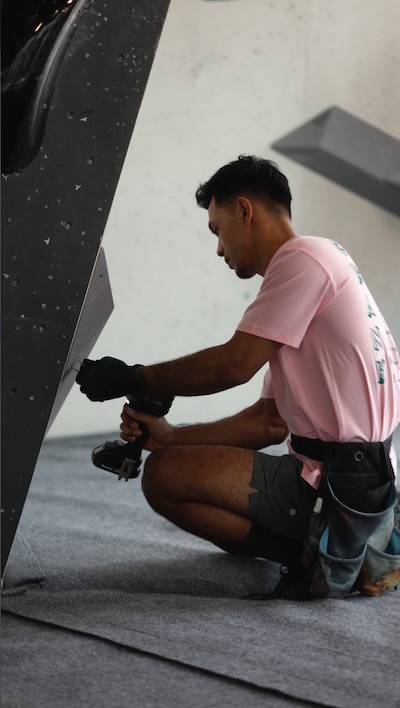 A climber grips a hold on an indoor climbing wall during a competition, illuminated by dramatic purple and blue lighting.